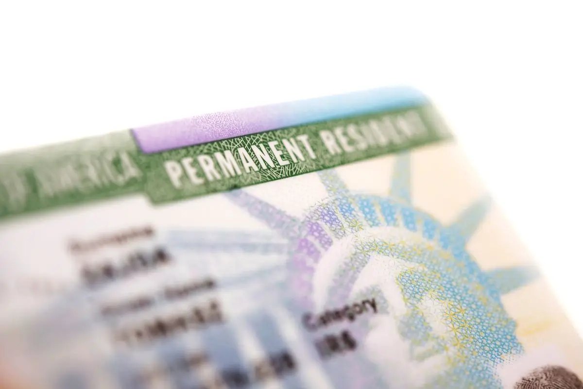 A close-up, shallow depth of field shot of a US Permanent Resident card, commonly known as a Green Card. The text "PERMANENT RESIDENT" is clearly visible along the top edge, with the image of the Statue of Liberty faintly in the background. The card is angled, drawing focus to its official details. The image symbolizes legal residency, immigration, and the path to living in the United States.