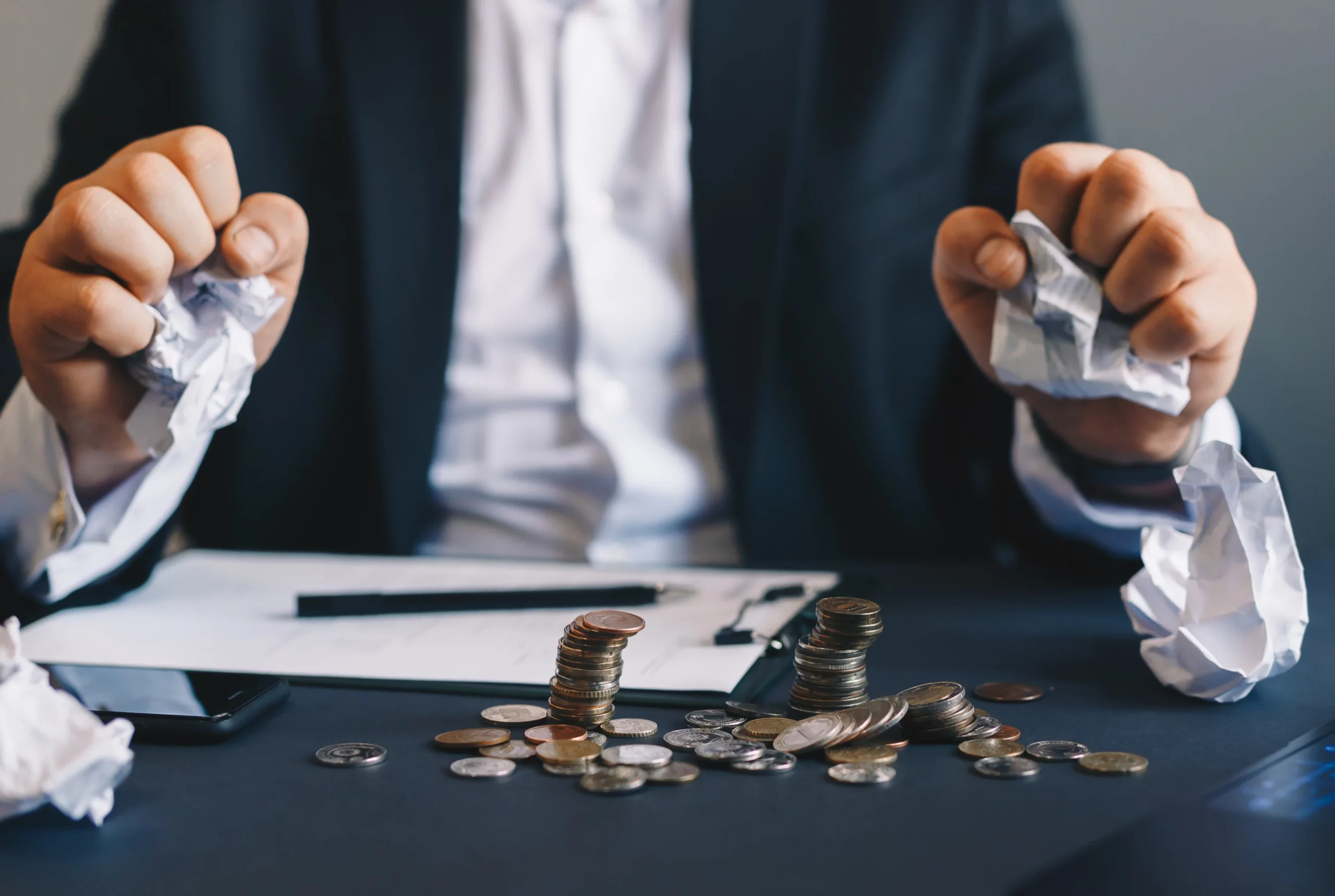 Frustrated businessperson clutching crumpled paper with cash on desk, showing financial stress.