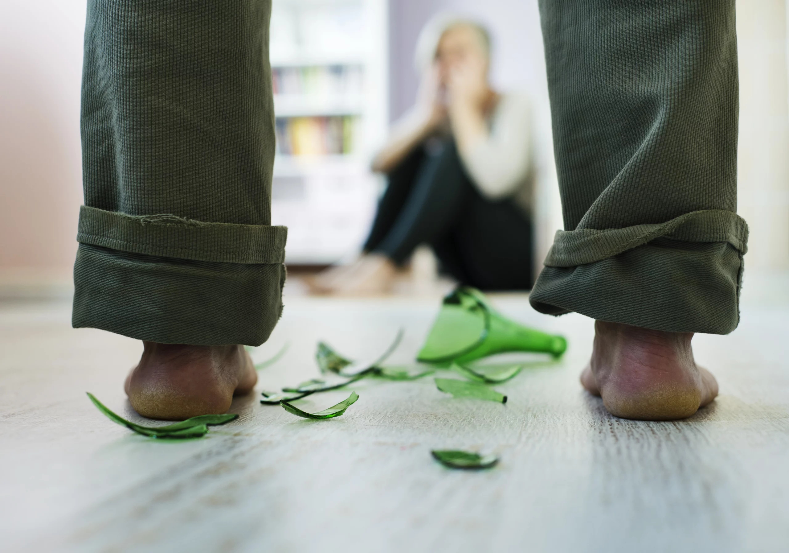 Bare feet approaching broken green glass on a floor, with a blurred seated person in the background—symbolizing danger, trauma, and vulnerability of domestic-violence victims.